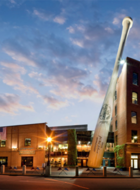 This image shows a visitor holding a Louisville Slugger bat at the Louisville Slugger Museum, with an exhibit in the background about the famous bats used by legendary baseball players.