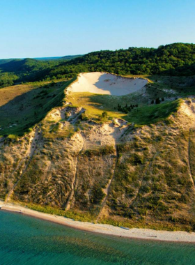 This image shows the towering dunes at Sleeping Bear Dunes National Park, with visitors hiking up the sandy hills, overlooking Lake Michigan’s stunning shoreline and forested landscapes.

