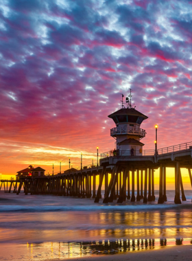 This image shows surfers riding waves at Huntington Beach, also known as Surf City USA. The scene captures surfers balancing on their boards as they glide across blue ocean waves. The sandy beach stretches in the background, lined with umbrellas and sunbathers enjoying the day. The iconic Huntington Beach Pier stands tall, with people watching surfers and soaking in the lively surf culture. The image reflects the vibrant energy of the beach.