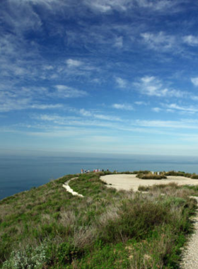 This image shows hikers walking along a scenic trail in Laguna Coast Wilderness Park. The trail winds through lush green hills and offers panoramic views of the coastline and valleys below. The hikers, dressed in outdoor attire, carry backpacks and walking sticks. The clear blue sky and sunlight add to the vibrant beauty of the natural surroundings. The image highlights the peaceful and rewarding experience of hiking in Orange County.