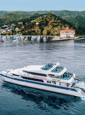 This image shows a ferry traveling across calm ocean waters toward Catalina Island. Passengers on the ferry deck are enjoying the fresh sea breeze and taking in views of the blue Pacific Ocean. The island, with its green hills and coastline, becomes visible in the distance. Seagulls fly above the water, adding to the sense of adventure. The image perfectly captures the anticipation and beauty of traveling to Catalina Island.