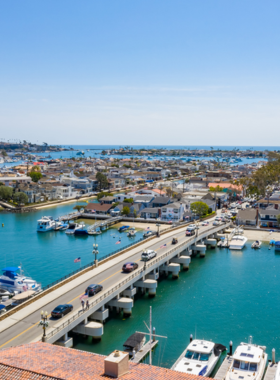 This image shows people walking along the waterfront at Balboa Island. The scene includes calm harbor waters reflecting boats and charming coastal homes. Visitors stroll leisurely, some carrying ice cream cones, while others sit on benches enjoying the view. Colorful shops and restaurants line the path, creating a small-town coastal vibe. The peaceful environment, with boats gently bobbing in the water, captures the relaxed charm of Balboa Island.