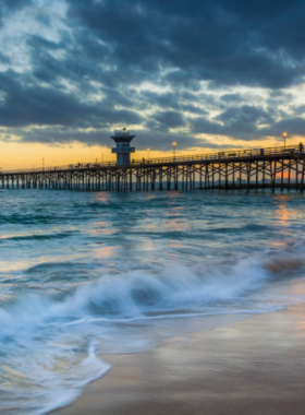 This image shows the Seal Beach Pier at sunset, with the sky painted in shades of orange, pink, and purple. The calm ocean reflects the colors of the setting sun, while visitors walk along the wooden pier. Silhouettes of people can be seen relaxing and enjoying the view. The golden sand and gentle waves create a peaceful coastal scene. The image highlights the beauty and tranquility of watching a sunset at Seal Beach.