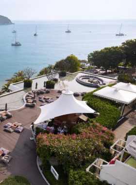 This image shows The Nai Harn Resort, perched above the Andaman Sea. It showcases guests enjoying rooftop drinks and locally-sourced meals while the resort emphasizes sustainability with its eco-conscious initiatives and stunning sea views.