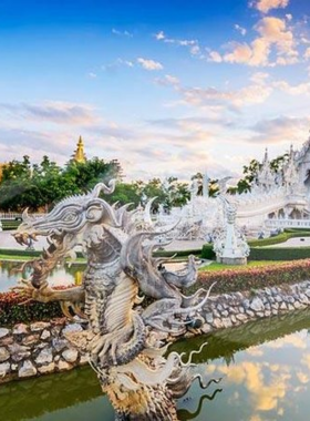 This image shows the iconic White Temple of Chiang Rai with its intricate white façade shimmering under the sunlight. The reflective pond in front of the temple adds to its ethereal beauty. The bridge leading to the temple is lined with detailed sculptures, representing the journey from worldly desires to enlightenment. The serene blue sky in the background enhances the temple’s stunning architectural design.