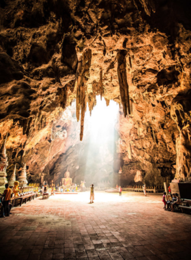 This image shows explorers entering the Tham Khao Luang Cave in Phetchaburi, surrounded by magnificent stalactites and stalagmites, and ancient Buddhist shrines.