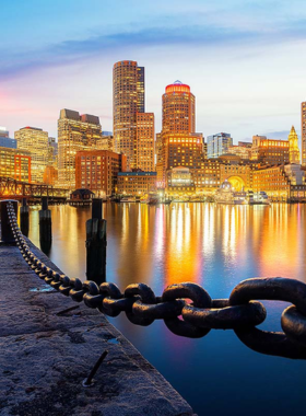 This image shows Boston Harbor at sunset, with calm waters reflecting the warm colors of the sky. Boats are docked along the waterfront, and visitors are strolling by, enjoying the serene views and the harbor’s rich maritime history.