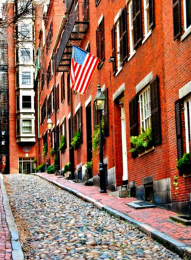 This image shows the charming streets of Beacon Hill in Boston, with its historic cobblestone roads, gas-lit lamps, and Federal-style townhouses. Visitors are walking along picturesque Acorn Street, often called one of the most beautiful streets in America.