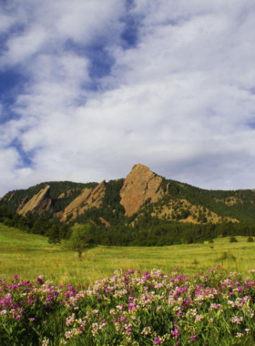 "This image shows the stunning Flatirons rock formations in Boulder, Colorado, with hikers exploring the trails beneath the towering peaks. The landscape offers panoramic mountain views, clear skies, and a serene environment, making it a perfect destination for outdoor enthusiasts."