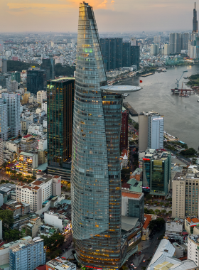 This image shows a breathtaking panoramic view from the Bitexco Saigon Skydeck, with the Saigon River and city skyline stretching into the distance. Tall skyscrapers and historical buildings create a stunning contrast, showcasing the modern development alongside traditional architecture. Visitors can enjoy the stunning 360-degree view of Ho Chi Minh City, with the sunset providing a dramatic backdrop to the urban landscape below, making it an unforgettable sightseeing experience.