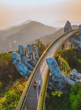 This image shows the iconic Golden Bridge at Ba Na Hills, nestled among the lush green mountains. The bridge, held up by giant stone hands, offers breathtaking views of the surrounding landscapes. Visitors are seen walking along the bridge, enjoying the fresh mountain air and stunning vistas. The beautiful gardens and hiking trails nearby add to the charm, making it a must-visit destination for nature and photography lovers.