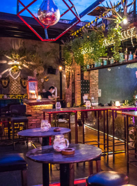 This image shows Broma: Not a Bar, a cozy rooftop bar in Ho Chi Minh City. The bar is lit with colorful fairy lights, creating a warm and casual atmosphere. People are seated on the terrace enjoying craft beers and cocktails while soaking in the city views. The setting includes wooden tables and greenery, adding a rustic touch to the ambiance. A musician is seen playing live music, adding to the charm of this lively yet relaxed venue.