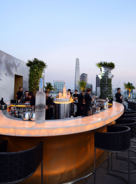 This image shows Glow Skybar, a trendy rooftop bar in Ho Chi Minh City with tropical vibes. The outdoor seating area has modern furniture surrounded by greenery. People are seen sipping cocktails under the open sky while enjoying the breathtaking city views. The ambiance is vibrant, with upbeat music playing in the background. The bar area is stylishly designed, creating a chic and inviting space for socializing or celebrating special occasions with friends.