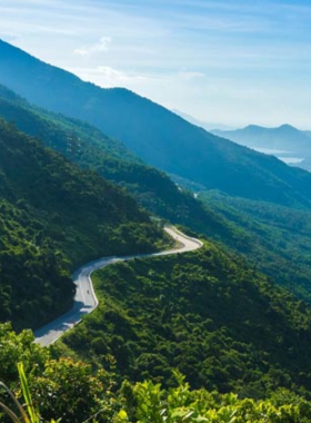 This image shows the iconic Hai Van Pass in Vietnam, a winding mountain road with stunning views of the coastline and surrounding greenery. The road snakes through mist-covered hills, offering a thrilling drive for adventurers and motorbike riders. The backdrop includes a clear blue sky, lush forests, and the sparkling ocean, making it a picturesque and unforgettable route.