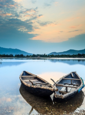 This image shows Lap An Lagoon in Hue, Vietnam, with its calm waters reflecting the surrounding mountains and sky. The peaceful setting features traditional fishing boats anchored along the shore, enhancing the local charm. The lagoon is framed by a backdrop of lush hills, creating a serene and picturesque scene perfect for nature photography and quiet escapes.