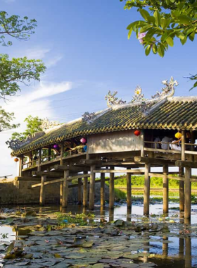 This image shows the picturesque Thanh Toan Bridge, a traditional Japanese-style covered bridge in a quiet village near Hue, Vietnam. The bridge is made of wood with intricate carvings and stands over a peaceful canal surrounded by lush greenery. It is a historic landmark and a gathering spot for locals. The image captures the rustic charm of the bridge and its surrounding area, which offers visitors a glimpse into authentic Vietnamese rural life and culture.