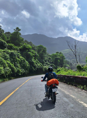 This image shows a traveler on a motorbike stopping at a scenic viewpoint during a Hidden Land Travel tour near Hue, Vietnam. The view includes a lush valley, distant hills, and a winding road. The traveler appears to be taking in the beauty of the landscape while the guide provides information about the area. The image highlights the adventurous and personalized experience of these tours, which explore lesser-known routes and offer an authentic glimpse of Vietnam’s countryside.