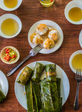 This image shows a variety of traditional Hue dishes, including bun bo Hue, banh beo, and nem lui, beautifully arranged on a table. The vibrant colors of fresh herbs, noodles, and sauces make the dishes look incredibly appetizing. A guide is explaining the history and preparation of the food to a group of eager participants. The bustling market and family-run eatery in the background add authenticity to the scene. The image captures the cultural and culinary richness of Hue’s local food tours.