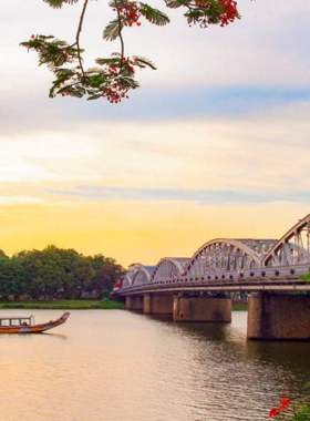 This image shows the calm waters of the Perfume River during sunset, with golden and orange tones filling the sky. The iconic Truong Tien Bridge is visible in the distance, its reflection shimmering on the water. A traditional dragon boat is gently floating, adding a cultural element to the scenic view. The image perfectly captures the serene and picturesque atmosphere of the Perfume River, making it a popular spot for peaceful boat rides and photography in Hue.