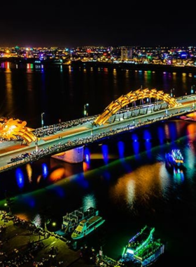 This image shows the iconic Dragon Bridge in Da Nang illuminated during its fire and water show. At night, the bridge breathes fire as part of a spectacular performance. The image captures the excitement of the crowd gathered along the banks, with street vendors selling snacks in the foreground. The vibrant lights and dramatic flames create a magical atmosphere, making this a must-see event for visitors to Da Nang.