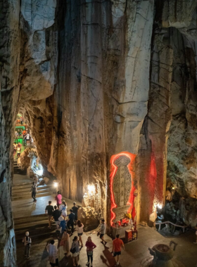 This image shows the breathtaking view from the top of the Marble Mountains in Da Nang. The image showcases the five limestone hills, with lush greenery and ancient pagodas scattered along the cliffs. A hiker stands at the summit, looking over the panoramic city view below. The spiritual and natural beauty of the location is highlighted by the Buddha statues inside the caves and the misty air that surrounds the area.