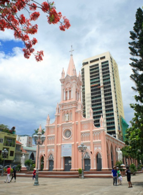 This image shows the beautiful Da Nang Cathedral, also known as the "Pink Church," with its rose-colored exterior and Gothic spire. The church stands out against the backdrop of the city’s skyline, showcasing French colonial architecture. The image captures the tranquil atmosphere of the church, with visitors walking around and taking photos. Its unique design makes it a photogenic and peaceful spot for cultural exploration in Da Nang.