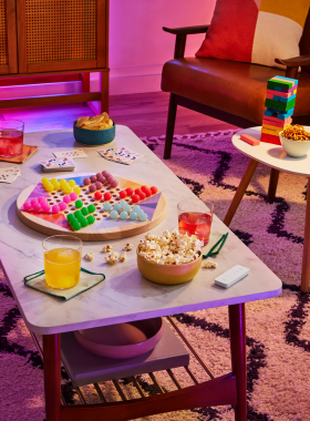 This image shows a group of friends sitting around a table, laughing and enjoying a game night. Board games, cards, and snacks like popcorn and drinks are scattered across the table. The scene is vibrant, with everyone smiling and engaged in friendly competition. The warm lighting and casual setting reflect the joy of spending quality time with friends, fostering connection and creating fun memories through an entertaining evening.