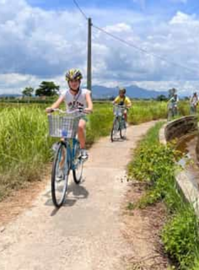 This image shows a cyclist riding along the scenic Cam Kim Bike Route in Hoi An. The route is surrounded by vibrant green rice paddies, calm riverbanks, and small traditional villages. A wooden bridge can be seen in the distance, adding a rustic charm to the peaceful setting. The cyclist appears relaxed, enjoying the fresh air and tranquil surroundings. This flat and beginner-friendly route is ideal for nature lovers and those looking to explore Hoi An’s countryside at a leisurely pace.