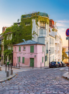 This image shows the charming streets of Montmartre, a historic and artistic neighborhood in Paris. The cobblestone pathways are lined with quaint cafés, galleries, and shops, offering a glimpse into the bohemian lifestyle that once thrived here. The image features the iconic Sacré-Cœur Basilica in the background, with its white dome standing prominently above the city. Montmartre is a beautiful district where visitors can enjoy a mix of culture, art, and picturesque views of Paris.