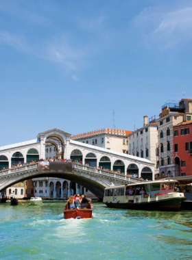 This image shows a serene gondola ride through the picturesque canals of Venice, Italy. The gondola glides beneath charming arched bridges, past pastel-colored historic buildings reflecting on the water. A gondolier, dressed in traditional attire, rows gracefully, creating a romantic and timeless experience in this enchanting city.