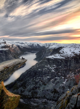 This image shows the dramatic rock formation Trolltunga, or "Troll’s Tongue," overlooking a breathtaking fjord in Norway. The cliff juts out over a vast landscape of blue waters and rugged mountains. Hikers stand at the edge, taking in the awe-inspiring view after completing the challenging 8-10 hour trek through Norway’s wilderness.