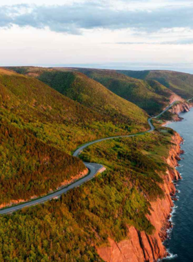 This image shows the scenic Cabot Trail, a winding coastal road that stretches along the cliffs of Cape Breton Island in Nova Scotia. The view from the road is breathtaking, with rolling green hills meeting the deep blue ocean. A car drives along the highway, surrounded by stunning landscapes, including dense forests and dramatic sea cliffs. The sky is clear, allowing the sun to shine on the road, making it perfect for a peaceful and picturesque drive. Along the way, travelers can stop at lookout points, hike nearby trails, or enjoy fresh seafood at charming coastal restaurants. The Cabot Trail is known as one of the most beautiful road trip destinations in the world, offering a mix of natural beauty and cultural charm. Whether you’re traveling solo, with friends, or on a family adventure, this drive is an unforgettable journey through Canada’s breathtaking East Coast scenery.