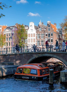 This image shows tourists on a canal cruise in Amsterdam, enjoying the scenic beauty of the city from the water. The iconic canals of Amsterdam, lined with beautiful historic buildings and charming bridges, offer a peaceful and unique way to see the city. The boat tour gives visitors a chance to learn about the rich history of Amsterdam while enjoying the picturesque views, with houseboats and lush greenery lining the canal banks.