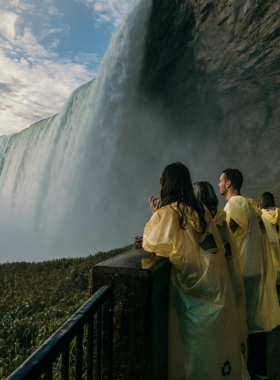 This image shows visitors on the Journey Behind the Falls tour, standing on observation platforms behind the massive Niagara Falls. They are surrounded by mist as they gaze at the powerful water cascading down from above. The image highlights the thrill of being so close to the falls, with the roar of the water creating a dramatic and unforgettable experience for those who take the tour."