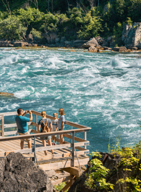 This image shows visitors walking along the White Water Walk, a scenic boardwalk that runs parallel to the raging Niagara River. Tourists can be seen gazing at the tumultuous rapids and swirling white waters that make this area famous. The lush green foliage and the powerful, fast-moving river create a thrilling and peaceful experience, giving visitors a close-up view of the wild, natural beauty of Niagara Falls."