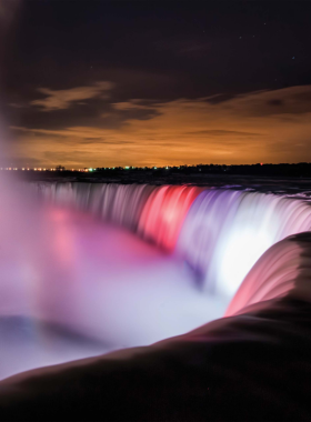 This image shows Niagara Falls illuminated at night, with vibrant, colorful lights casting a beautiful glow on the falling water. The falls are lit up in a rainbow of colors, creating a magical and surreal atmosphere. Tourists gather on the viewing platforms to witness this spectacular light show, which enhances the natural beauty of the falls and offers a unique experience for visitors in the evening."