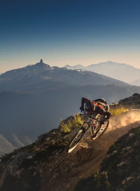 This image shows a mountain biker speeding down a forest trail in Whistler, surrounded by tall evergreen trees. The dirt path winds through the wilderness, creating an exciting adventure for thrill-seekers. The biker is wearing protective gear, navigating the rugged terrain with ease. The sunlight filtering through the trees adds a magical touch to the scene. The image captures the exhilarating experience of mountain biking in Whistler’s famous bike park, which attracts riders from all over the world.