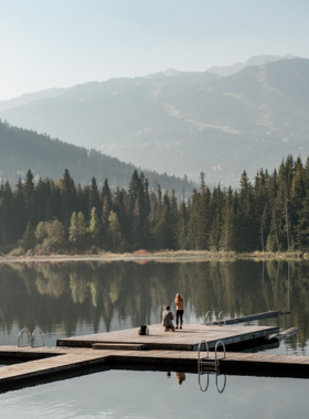 This image shows the serene beauty of Lost Lake, surrounded by lush green forests and reflecting the blue sky. A well-maintained hiking trail runs alongside the lake, where people are enjoying a peaceful walk and a cyclist is passing by. The calm waters create a mirror-like reflection of the towering trees. This image captures the tranquility of Lost Lake, a perfect spot for hiking, biking, and relaxing while soaking in the stunning natural scenery of Whistler.