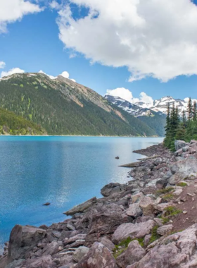 This image shows a breathtaking view of Garibaldi Lake, its turquoise waters surrounded by majestic snow-capped mountains. A well-marked hiking trail leads to this stunning destination, with hikers making their way up the rugged path. The dense green forest contrasts with the bright blue sky, creating a perfect nature escape. This image captures the rewarding beauty that awaits those who hike to Garibaldi Lake, one of Whistler’s most stunning and sought-after natural attractions.