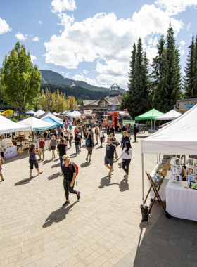 This image shows the vibrant Whistler Farmers Market, with colorful stalls displaying fresh produce, handmade crafts, and artisanal products. People browse the stalls, tasting samples and chatting with local vendors. The lively outdoor setting, surrounded by mountains, creates a warm and welcoming atmosphere. This image captures the essence of the market, where visitors can experience local flavors and unique handmade goods.