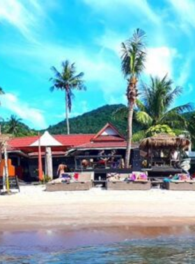 This image shows the relaxing beachfront atmosphere of Maya Beach Club in Koh Tao, where visitors enjoy cocktails and music under the open sky. The tropical setting includes the clear blue ocean and a sandy shoreline.