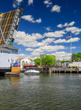 This image shows the Mystic River Bascule Bridge in Mystic, Connecticut, opening to let boats pass through. The iconic drawbridge lifts every hour, providing a fascinating engineering spectacle. The view of the counterweights pulling the bridge up and down is mesmerizing to watch. Visitors often grab a treat from nearby shops to enjoy while observing this unique landmark, adding an extra touch to the experience.