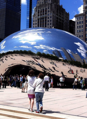 This image shows the famous Cloud Gate, also known as The Bean, in Millennium Park, Chicago. The large, shiny, bean-shaped sculpture reflects the city’s skyline, people, and the bright blue sky. Tourists are gathered around, taking selfies and admiring the unique structure. The surrounding park is lush and green, with pathways and benches where visitors can relax. The art piece creates a mesmerizing effect as it distorts and mirrors the scenery, making it one of the most photographed spots in Chicago.