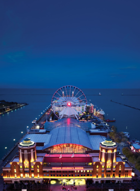 This image shows the iconic Centennial Wheel at Navy Pier in Chicago. The Ferris wheel stands tall against a bright blue sky, with enclosed glass gondolas offering stunning views of the city and Lake Michigan. Families, couples, and tourists are enjoying their ride, capturing breathtaking pictures from the top. The amusement park atmosphere is lively, with colorful lights and street performers. In the background, people are walking along the pier, visiting restaurants, and enjoying the waterfront experience.