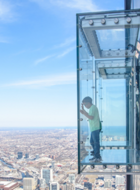 This image shows a breathtaking sunset view from the 360 Chicago Observation Deck, located on the 94th floor of the John Hancock Center. The golden-orange hues of the sunset cast a glow over the city’s skyline, while Lake Michigan stretches into the horizon. The glass windows reflect the twinkling city lights. Visitors are standing near the edge, looking out in awe, some capturing photos. The famous TILT experience, where visitors lean over the edge of the glass panel, adds a thrill to the scenic view.