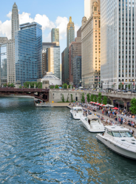 This image shows the beautiful Chicago Riverwalk, a pedestrian-friendly path along the Chicago River. People are walking, jogging, and sitting on benches while enjoying the waterfront view. Cafes, outdoor restaurants, and bars line the walkway, creating a lively atmosphere. The skyscrapers towering above reflect on the water, adding to the scenic charm. A tour boat is passing by, and a few people are kayaking in the river, making it an ideal place for relaxation and leisure.