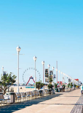 This image shows the iconic Ocean City Boardwalk, bustling with visitors enjoying the shops, arcades, and beach views. The wide boardwalk stretches along the coast, with colorful storefronts and signs, as people stroll by, taking in the lively atmosphere. Families walk past ice cream stands, while others stop for a moment to admire the scenic ocean views. The famous boardwalk has been a historical landmark in Ocean City since 1902, offering a fun and nostalgic experience for all ages.