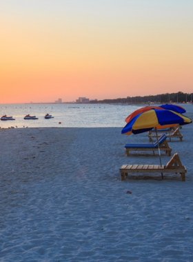 This image shows a beautiful view of Biloxi Beach during sunset. The sky is painted with warm shades of pink, orange, and purple, reflecting on the calm Gulf waters. People are walking along the soft white sand, some dipping their feet in the shallow waves while others relax on beach chairs under colorful umbrellas. A jet ski can be seen speeding across the water, leaving behind a white foamy trail. The peaceful atmosphere and breathtaking scenery capture the essence of a perfect day by the coast.