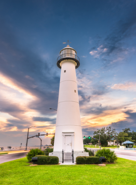 This image shows the historic Biloxi Lighthouse standing tall against a bright blue sky with white clouds. The white cast-iron structure has a classic black lantern room at the top, offering a panoramic view of the coastline. A group of tourists is gathered at the base, listening to a guide share fascinating stories about its history. The surrounding area features lush green grass, a paved walkway, and a scenic ocean view, making it a perfect spot for visitors interested in history and coastal beauty.