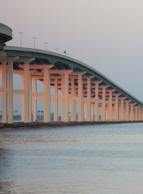 This image shows the Biloxi Bay Bridge stretching across the water with a pedestrian walkway alongside the road. People are seen walking and jogging, enjoying the fresh air and scenic view of the bay. The sky is partly cloudy, casting soft shadows on the bridge. In the distance, the city skyline and ocean waves create a peaceful backdrop. The bridge has safety railings and distance markers, making it an ideal spot for fitness enthusiasts and those looking for a relaxing stroll with a breathtaking view.